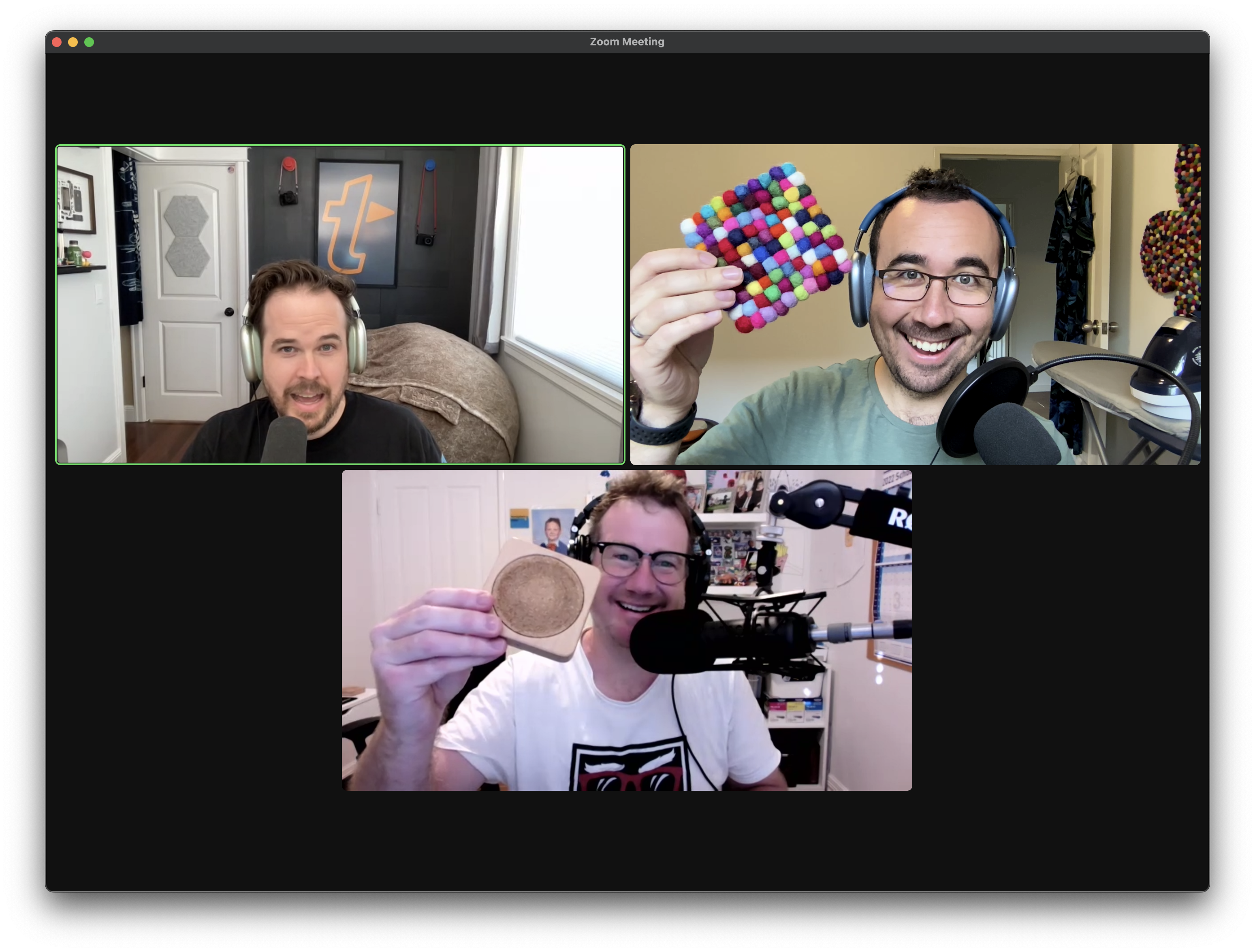 A Zoom call window with three men wearing headphones and showing drink coasters to their cameras