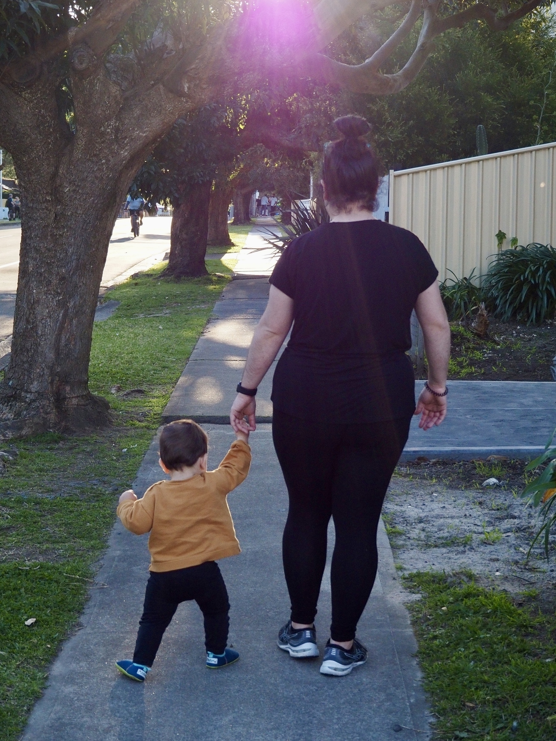 A mother and young child hold hands while walking along a footpath, with their backs to the camera.