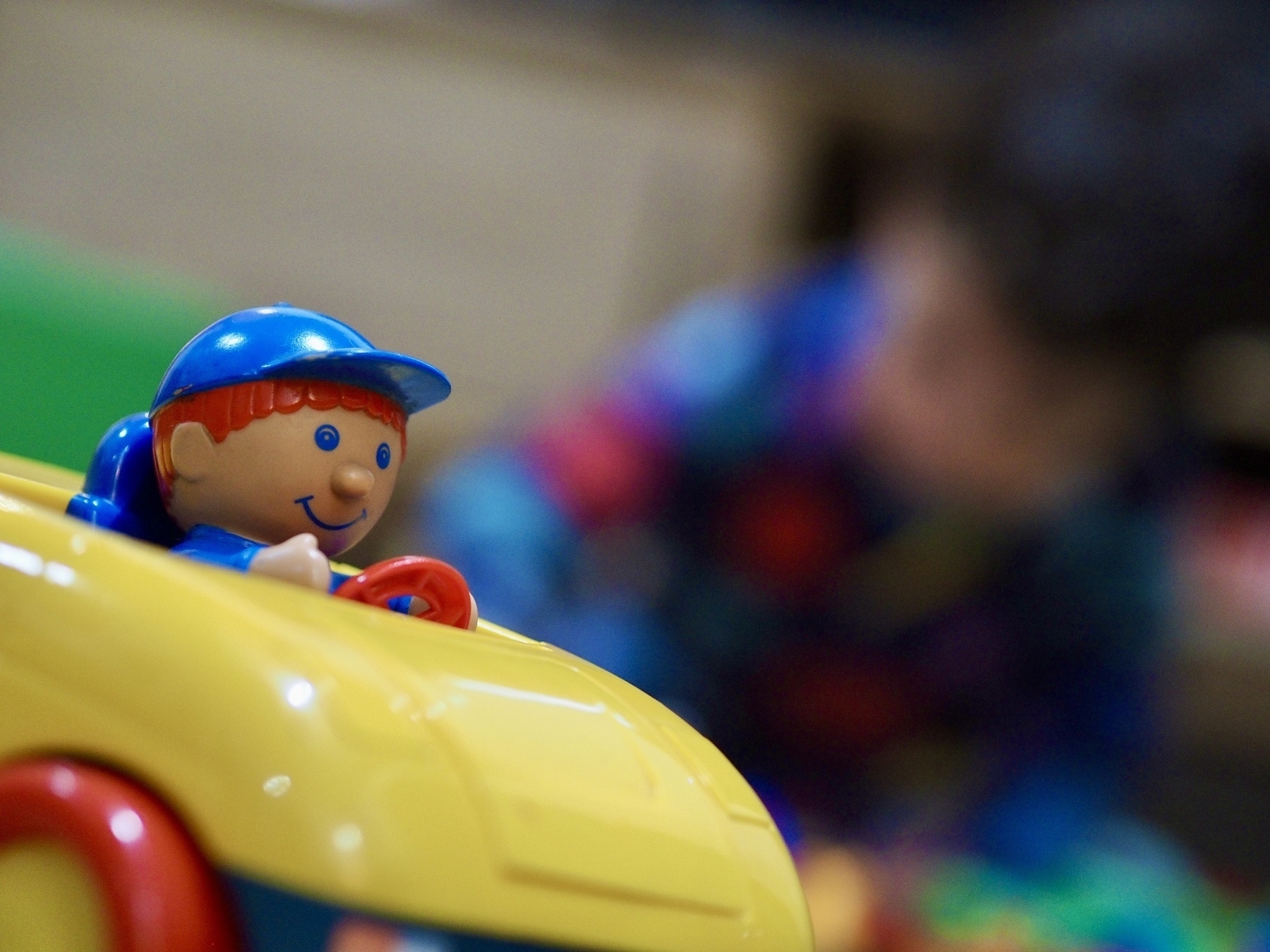 A close-up of a plastic figurine in a toy car with a blurred toddler in the background