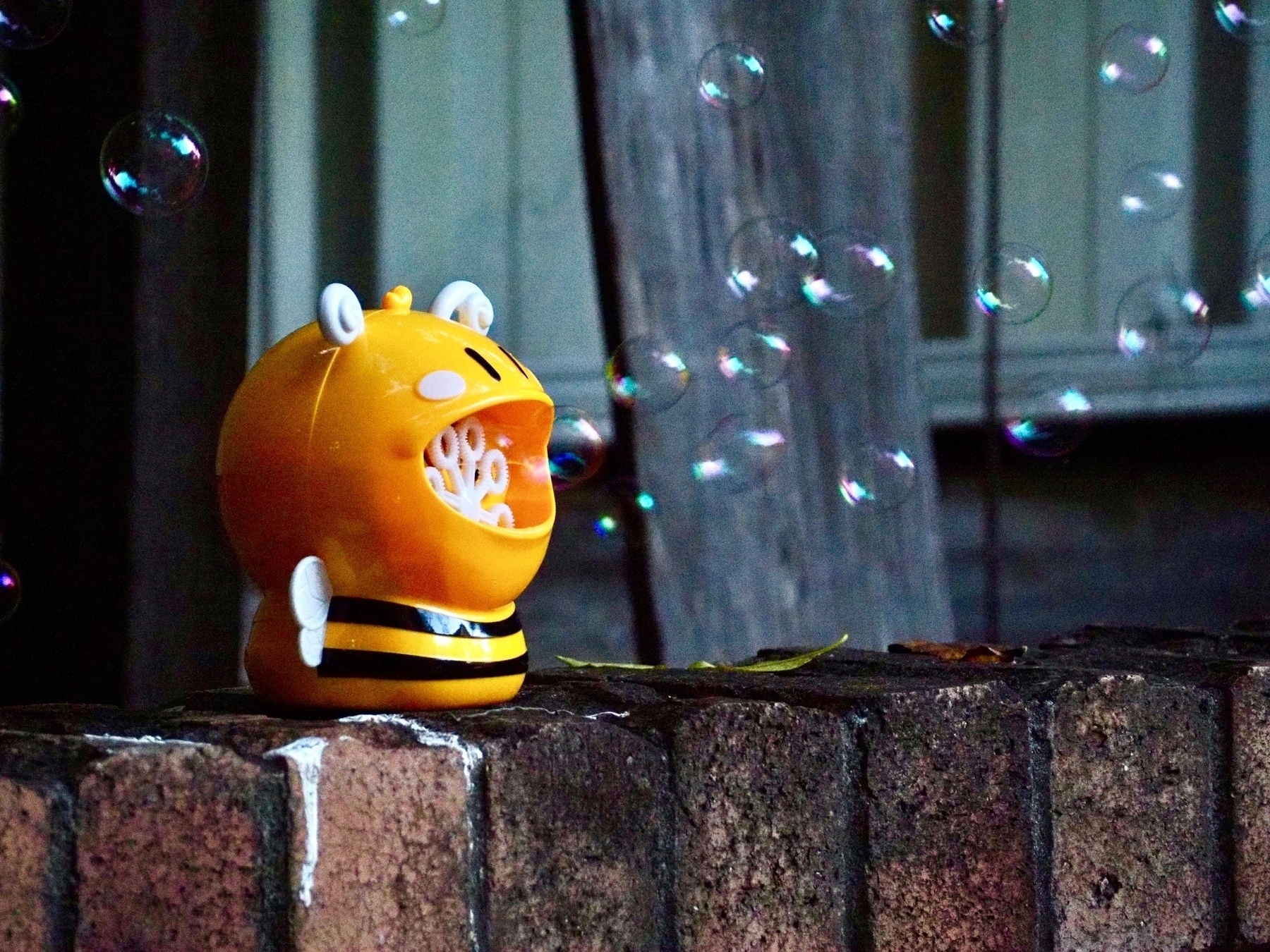 A yellow bubble machine in the shape of a bee sits on a brick retaining wall and spits bubbles out of its mouth.
