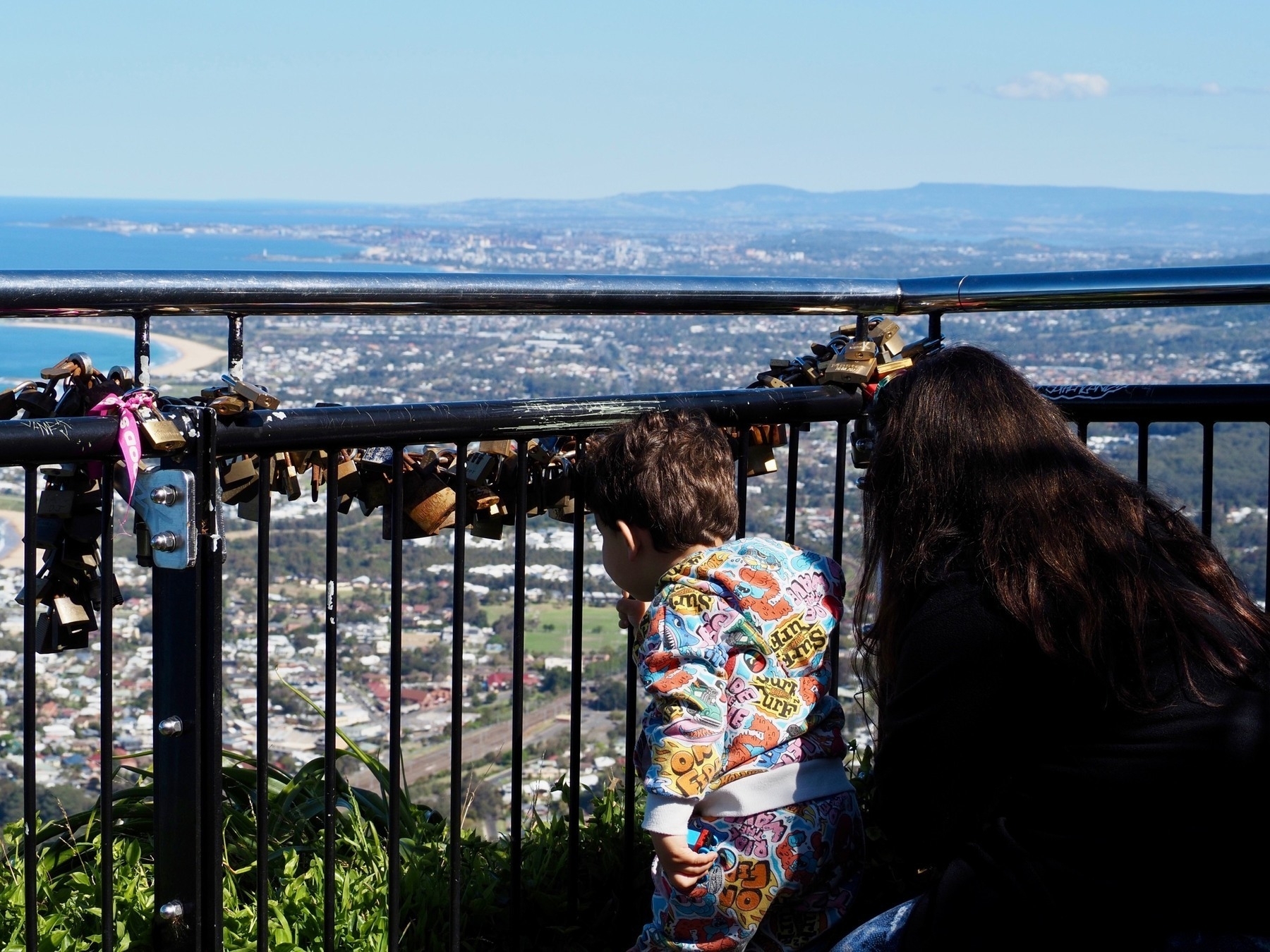 A mother and son look through a metal fence covered in locks over a a city and the sea.