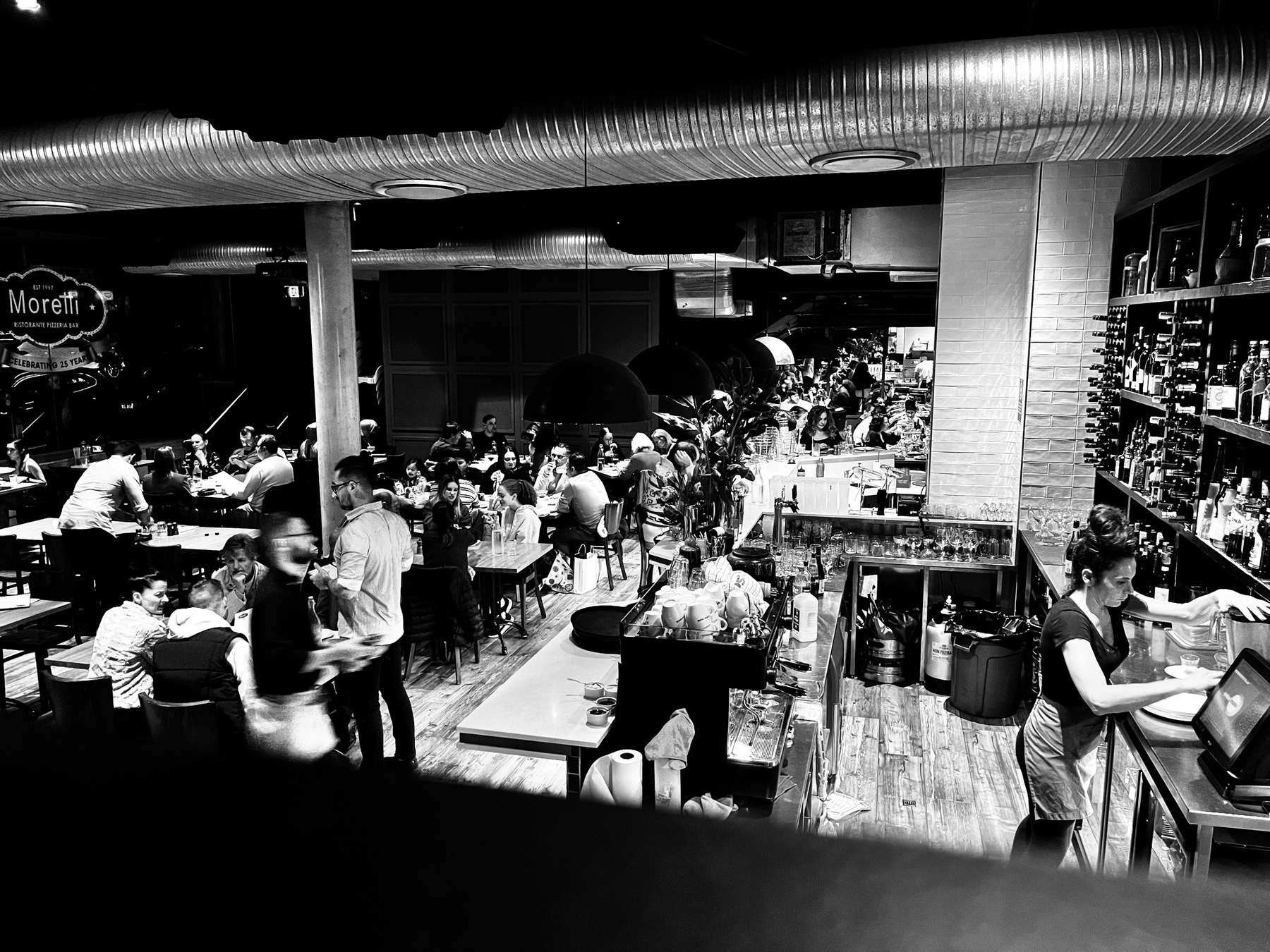A black-and-white image of a busy restaurant and bar, viewed from the upper level