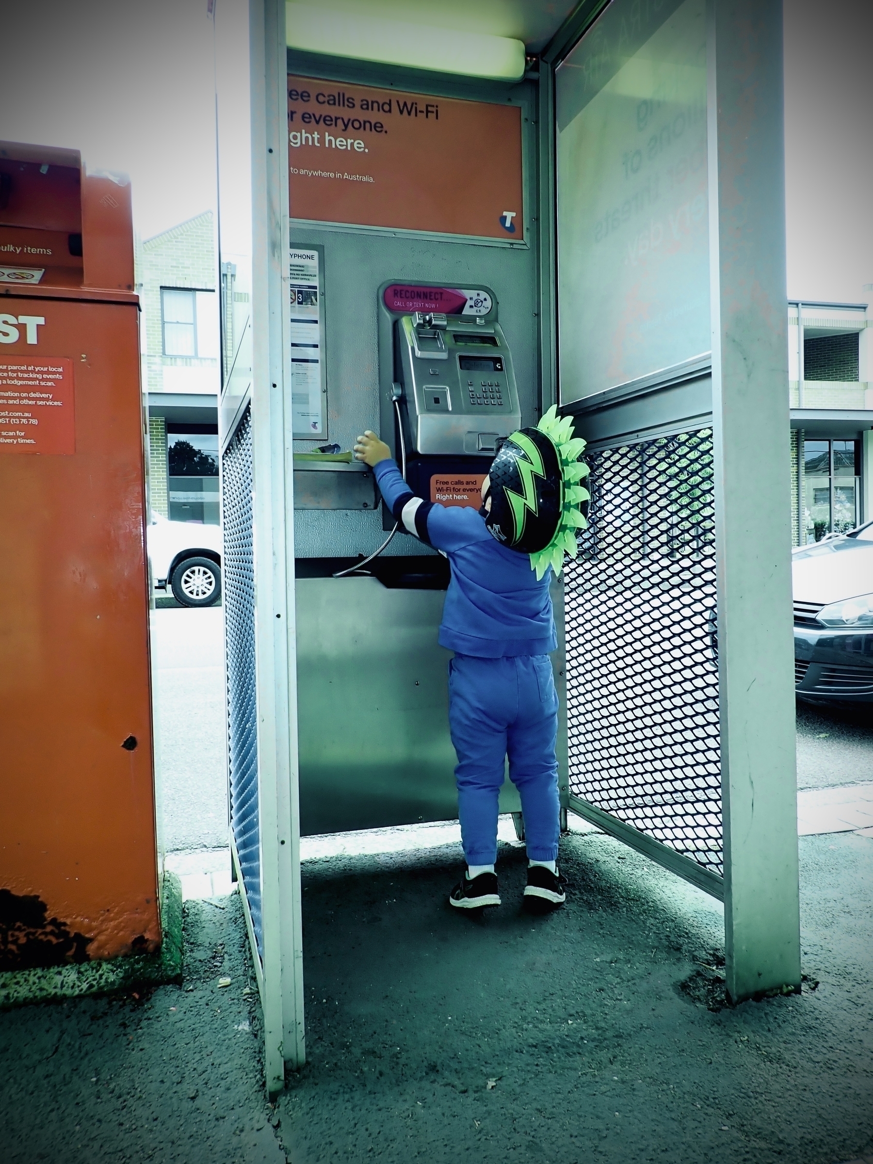 A young boy wears a spiky, green helmet in a phone booth.