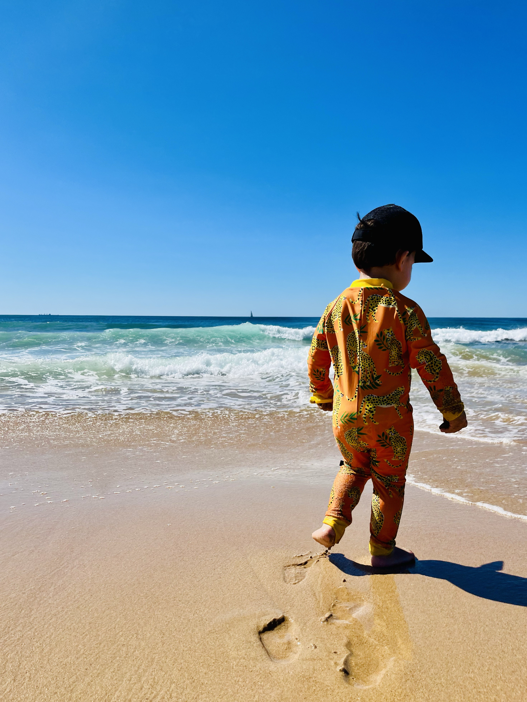 A toddler stands on sand in front of waves, under a cloudless sky.