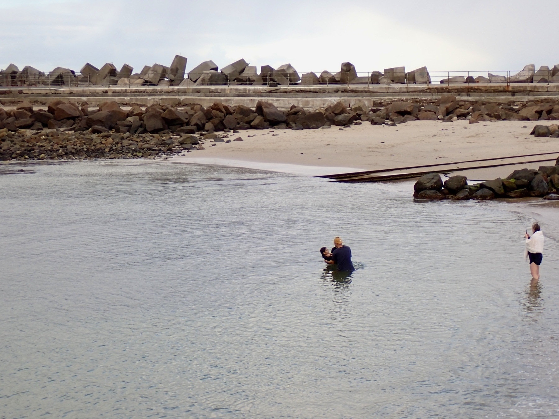 A person is dunked in water at a sheltered beach, with a rock wall in the background.