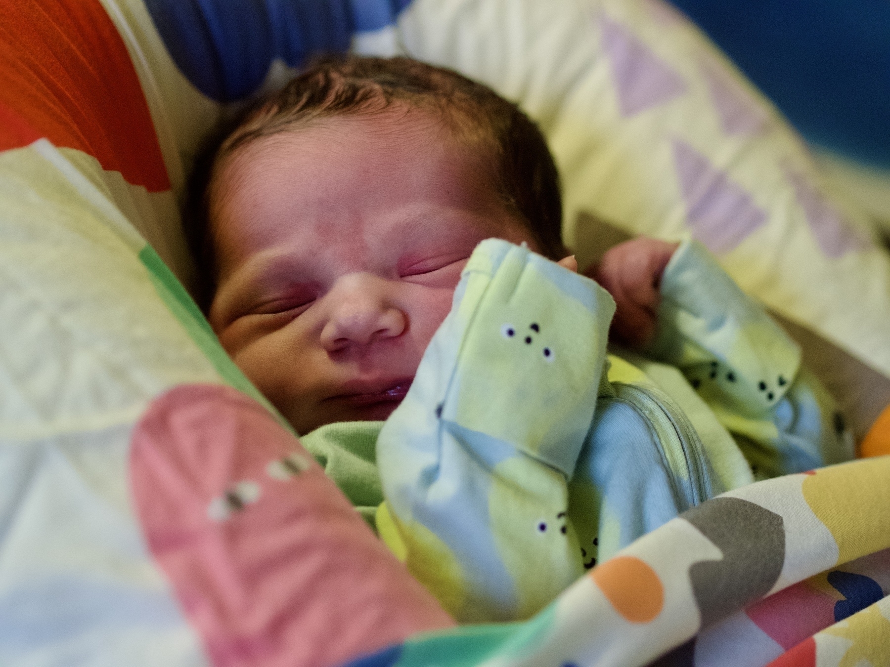 A newborn baby girl sleeps in a portable infant’s bed, while wearing pyjamas with a pattern of smiling pears.