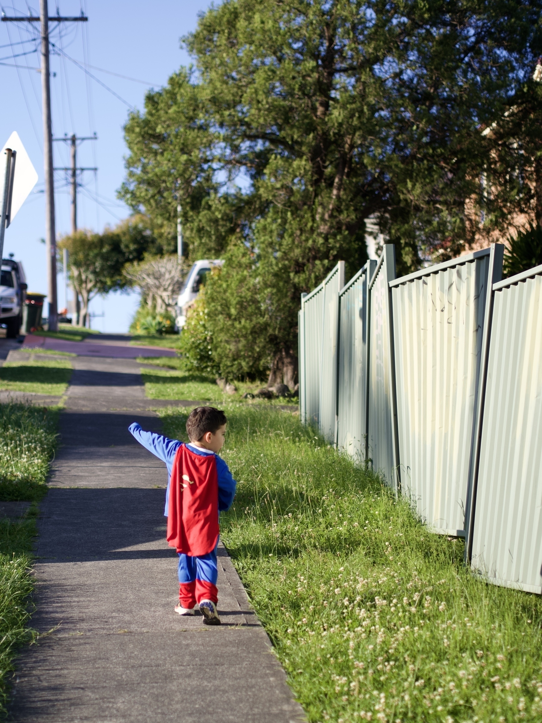 A young boy dressed as Superman runs up a footpath on a hill, with his left arm reaching upwards.