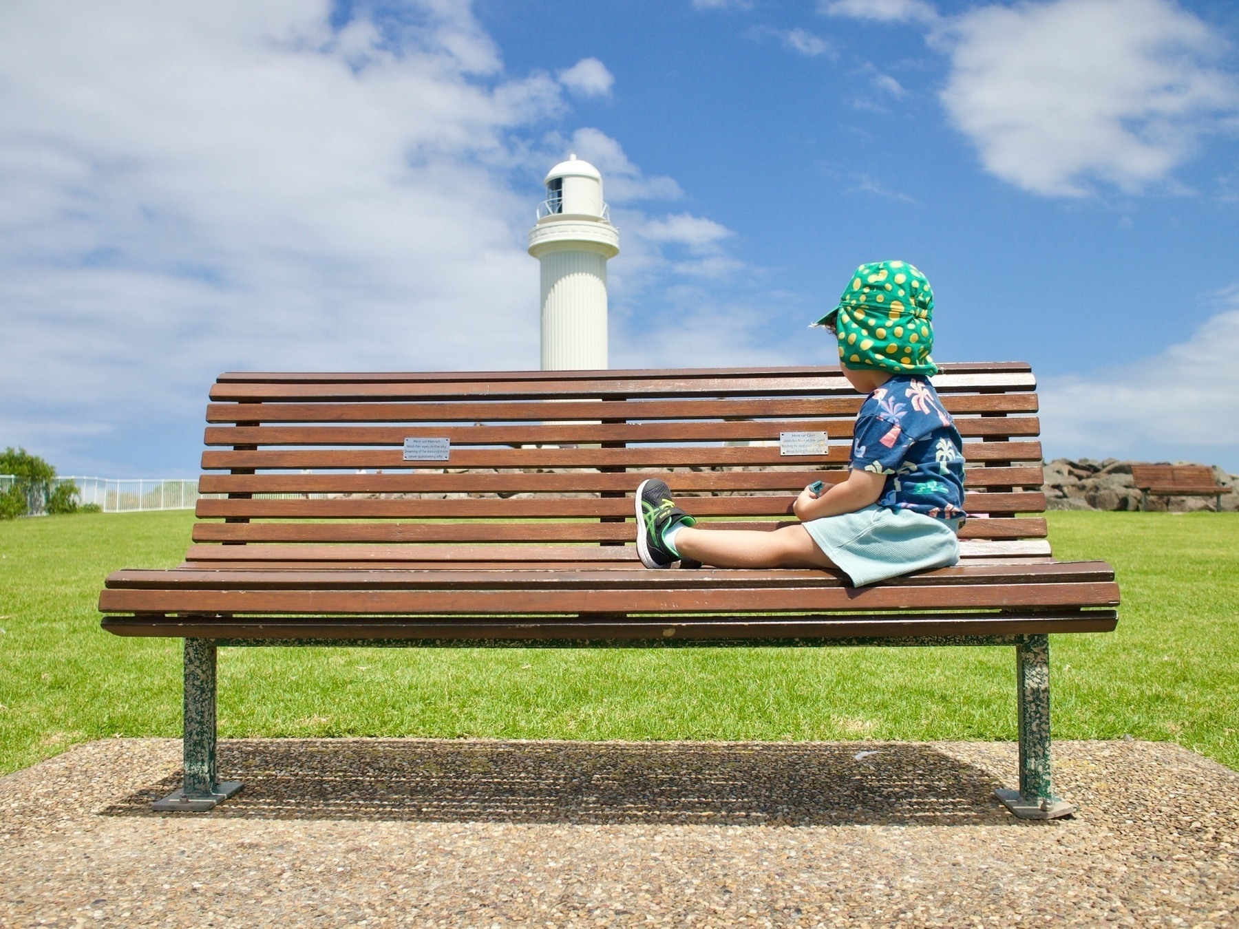 A toddler with a Dorothy the Dinosaur hat sits on a park bench in front of the lighthouse on Flagstaff Hill.