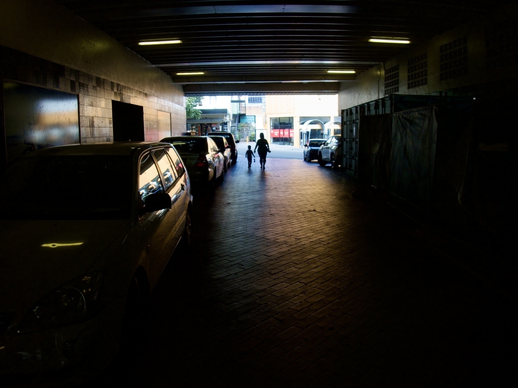 Silhouettes of a mother and son walking out of a dark tunnel filled with cars, towards a sunlit cul-de-sac