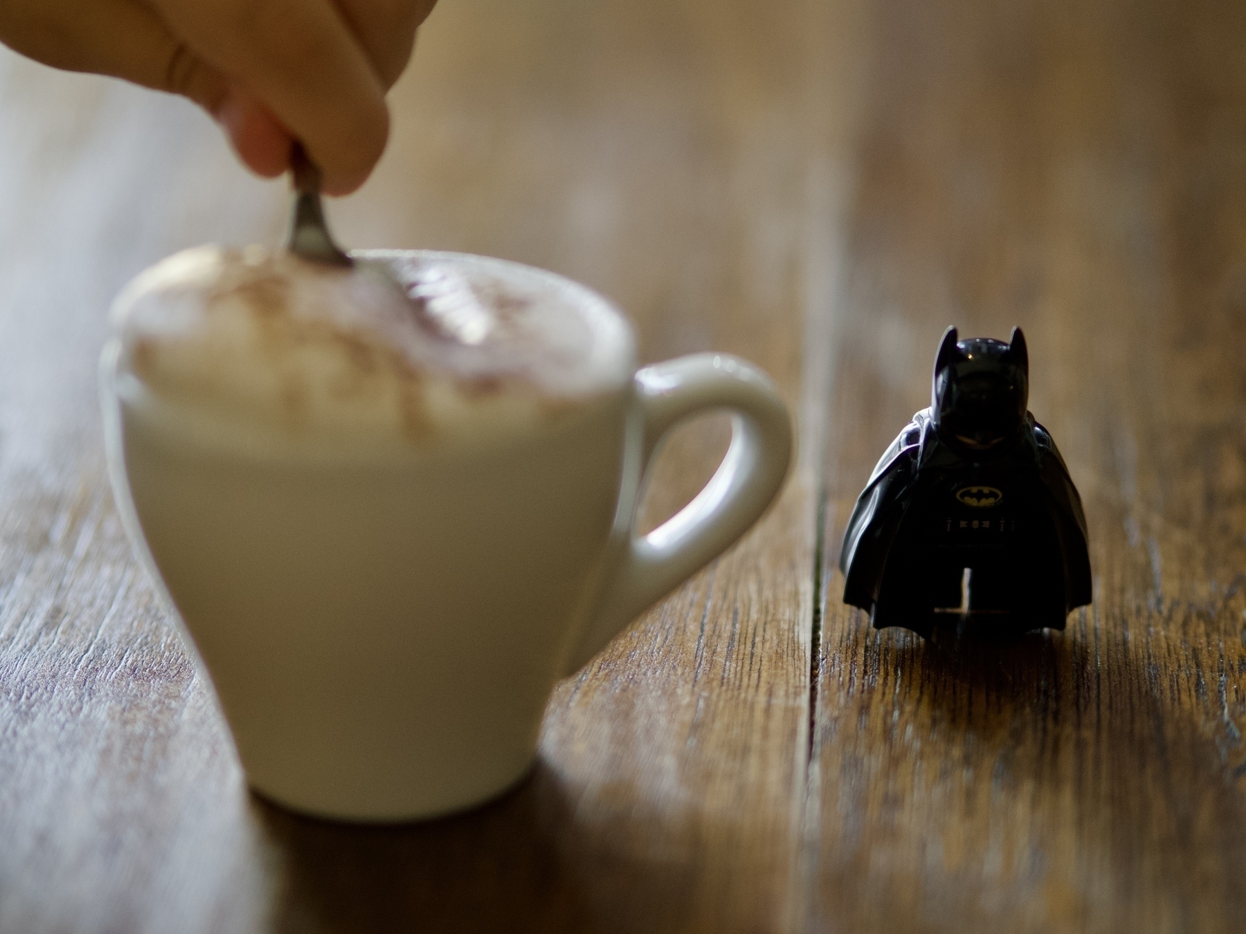A toddler's hand with a spoon above a small cup with froth, next to a Lego Batman figure on a wooden table