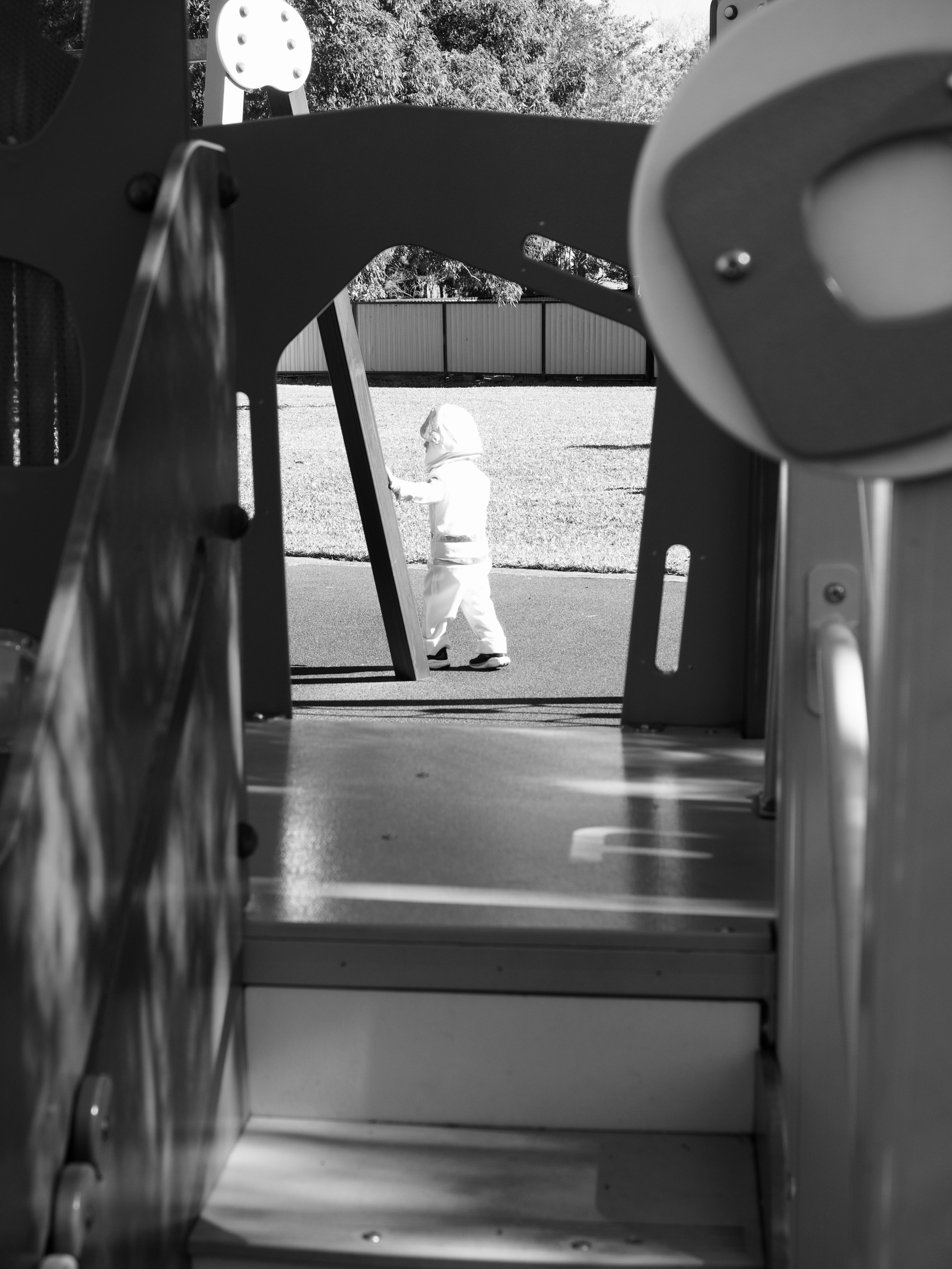 A black-and-white image, looking through play equipment at a young boy in an astronaut costume walking through a park