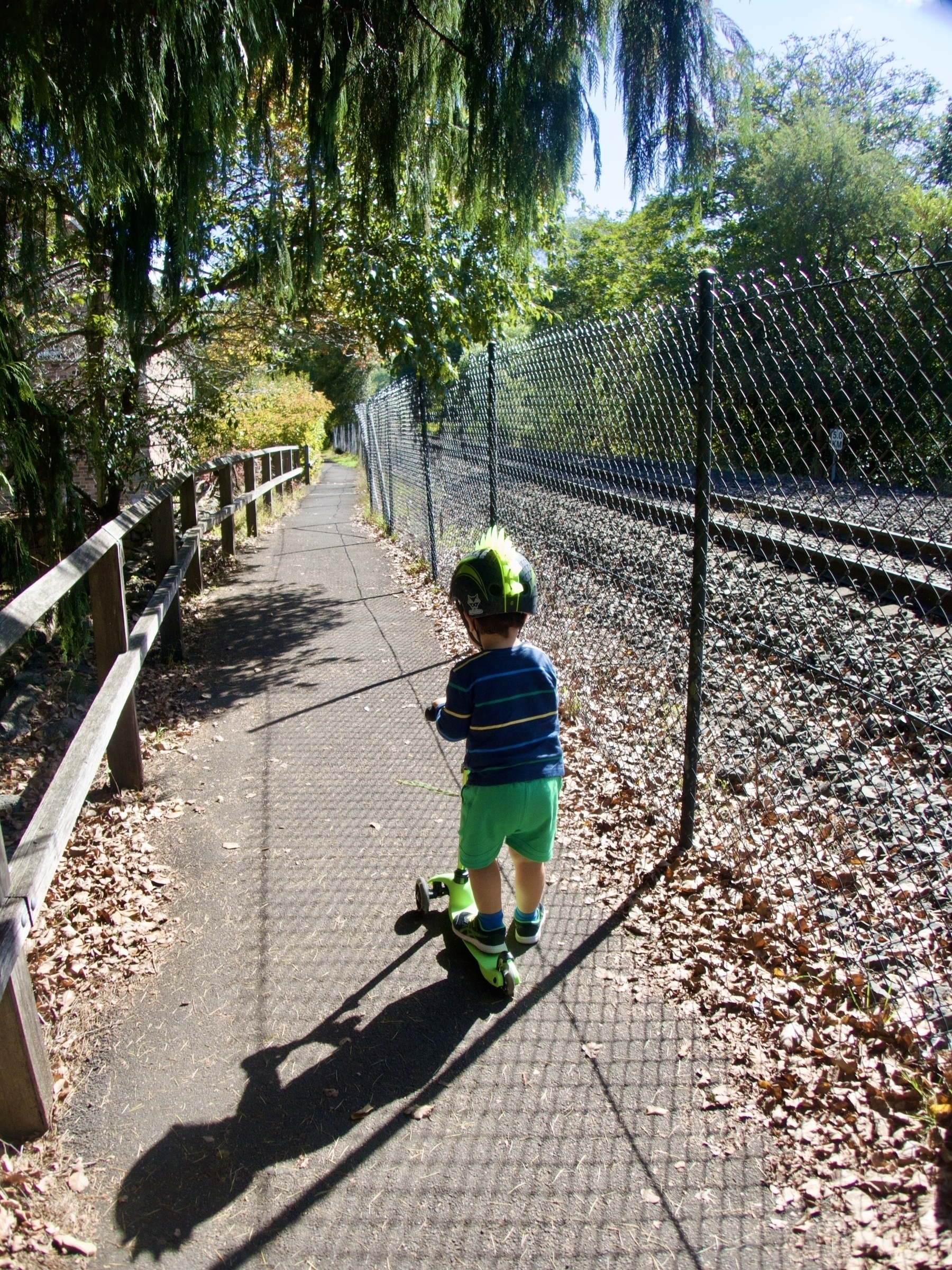 A kid seen from behind, riding a scooter while wearing a spiky green helmet, next to a railway with overhanging tree branches