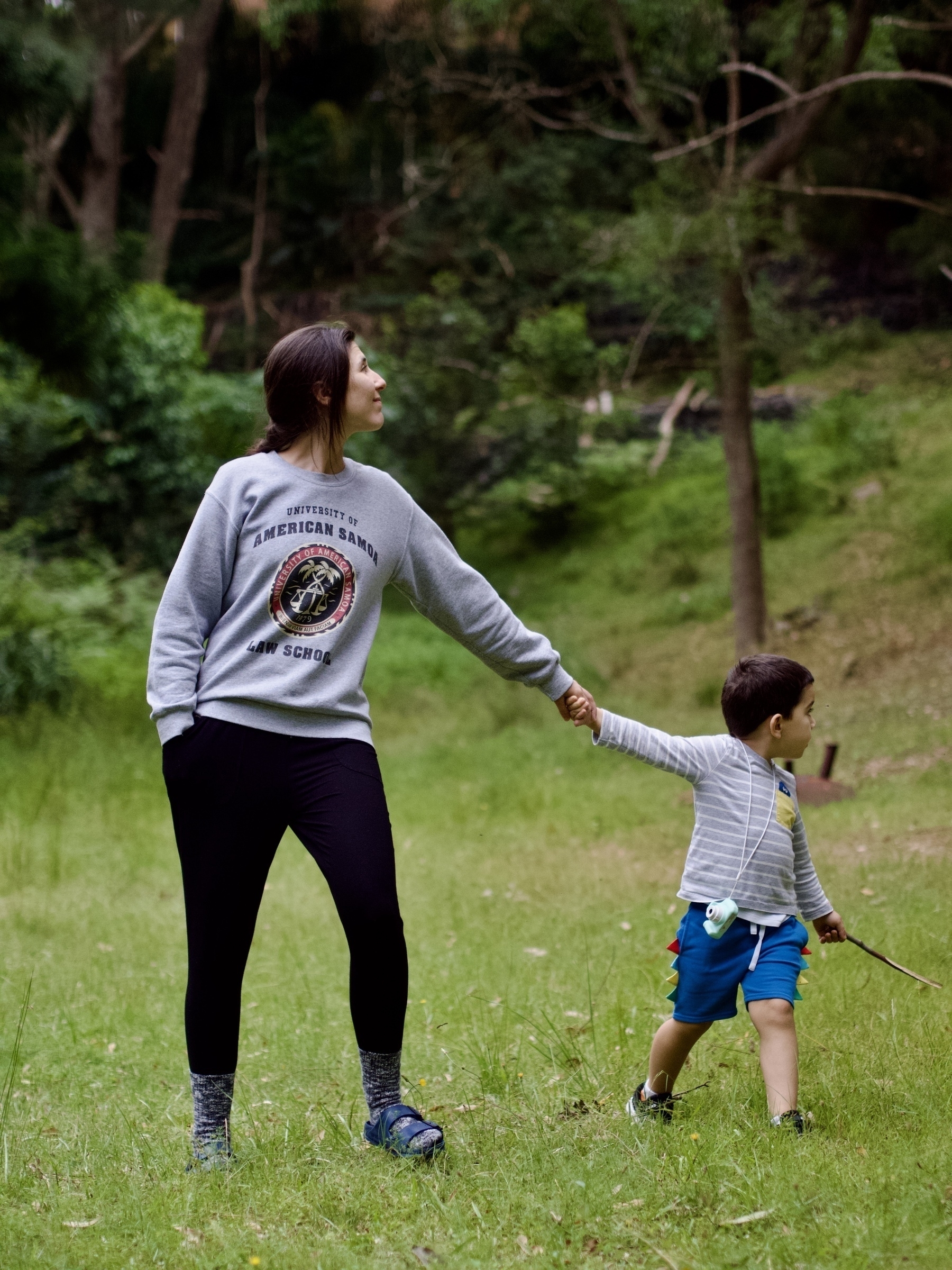 A young boy with a stick and a kid’s camera walks with a young woman in a ‘Better Call Saul’ top in a grassy clearing.