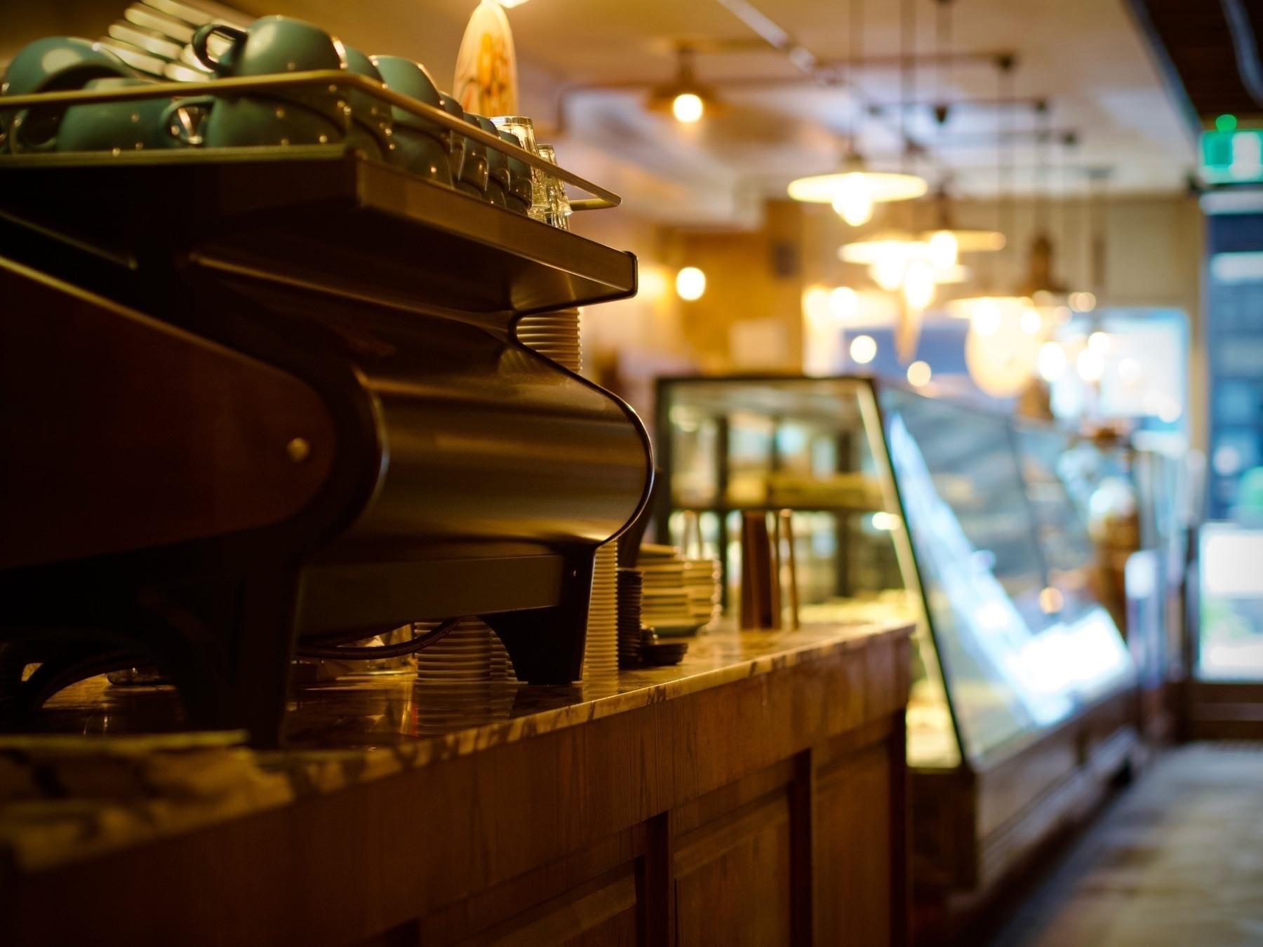 A large, brown coffee machine in the left foreground, extending past blurred café food cabinets to a doorway in the background