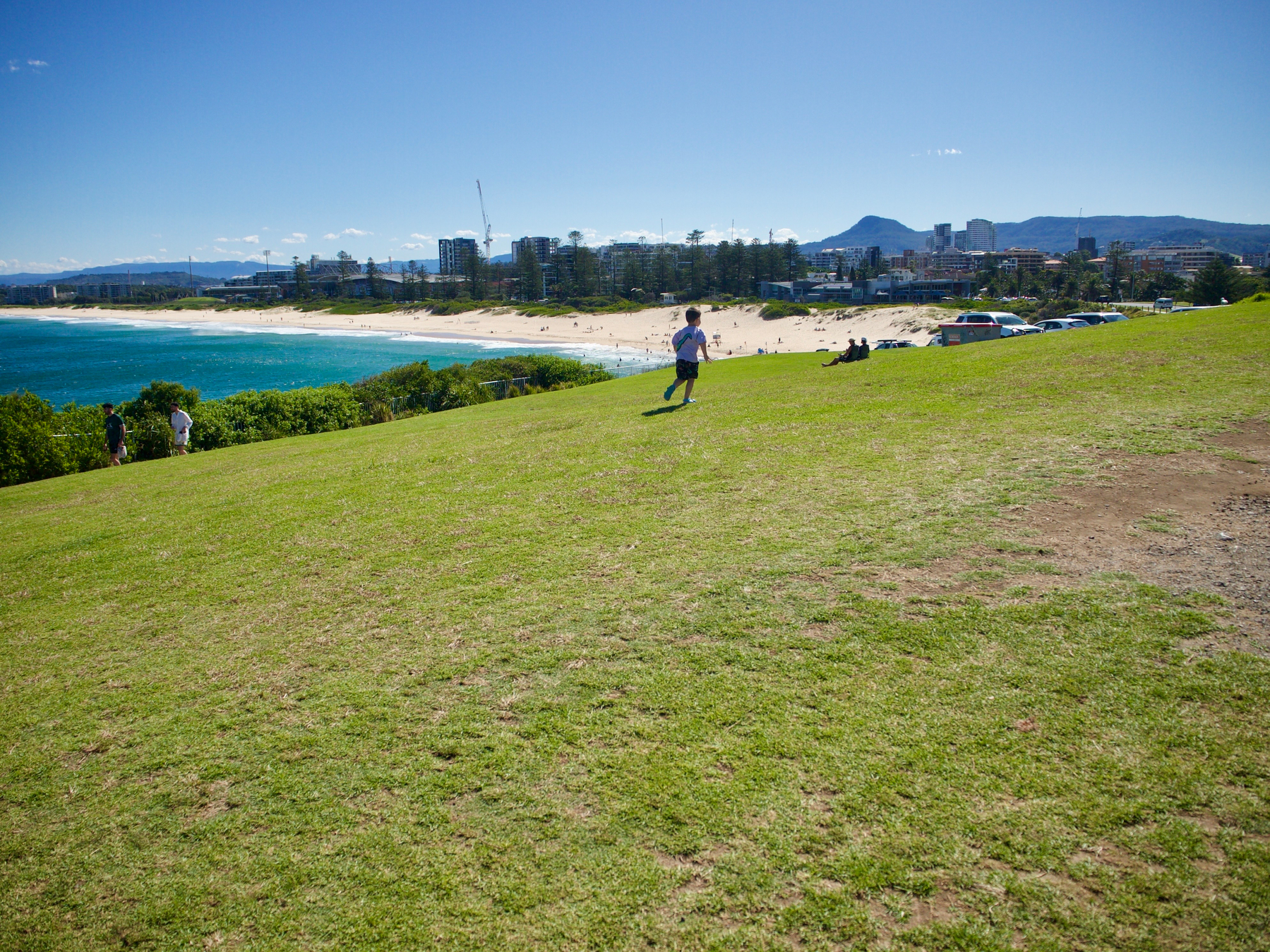 A child runs gleefully on a grassy hill (called Flagstaff Hill) with City Beach, buildings in Wollongong and the escarpment in the distance.