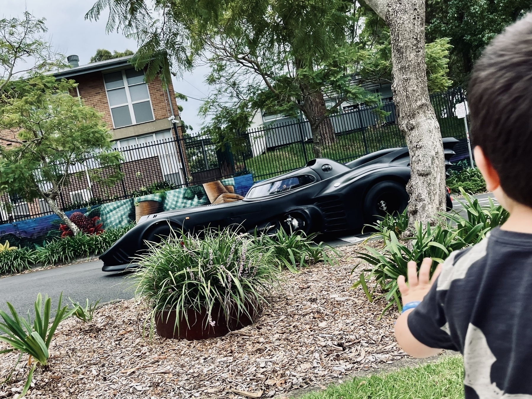 A child waves at a replica 1989 Batmobile driving through a leafy suburb.