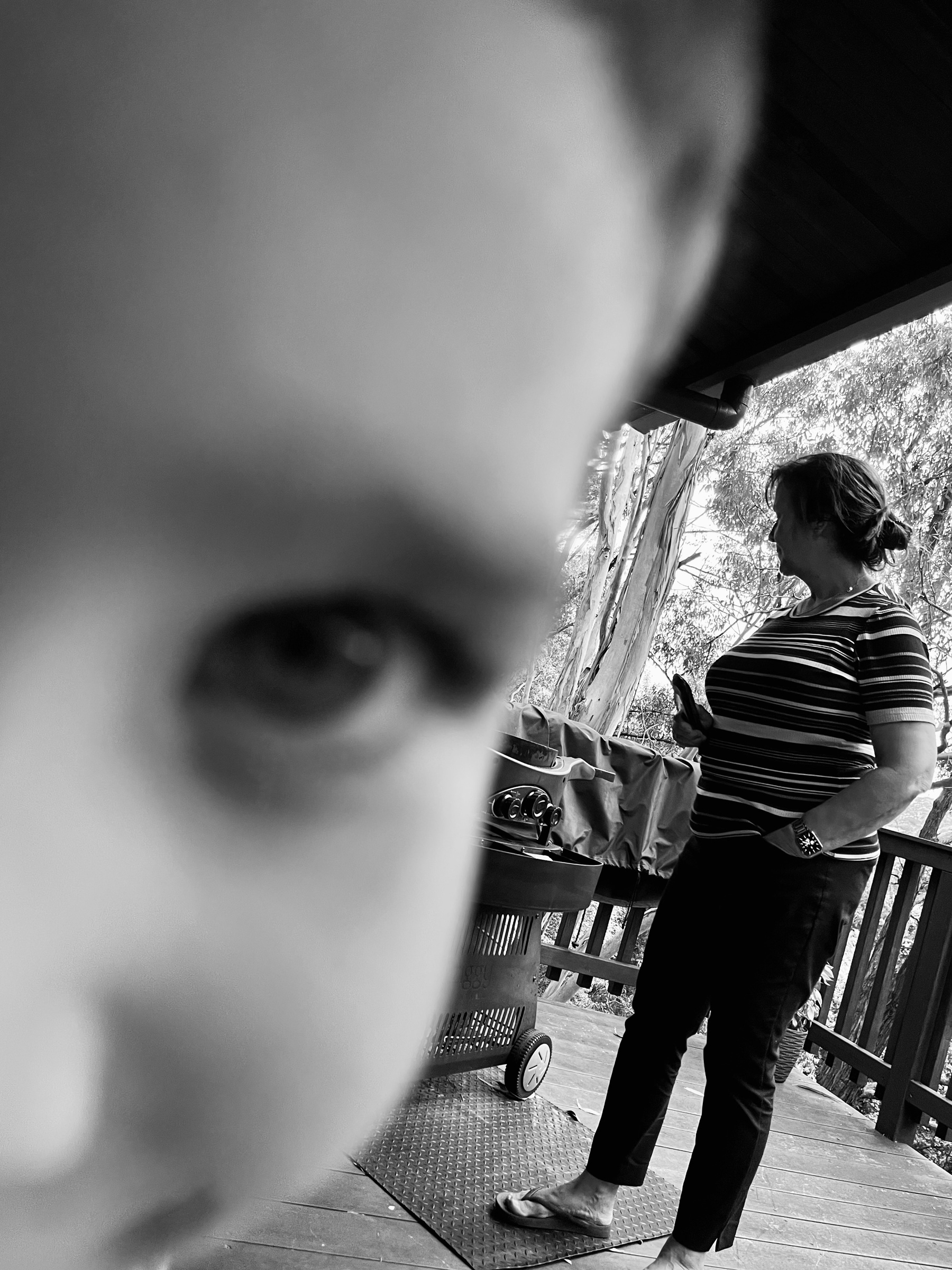 A black-and-white photo of a child’s face close to  the camera and a woman cooking meat on a barbecue with trees in the background